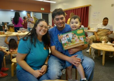 A mother, father, and smiling toddler pose together at a community event; the child holds a boxed kids' kitchen set.