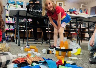 A young child in a superhero costume builds a tower from bottle caps and containers on a library floor.