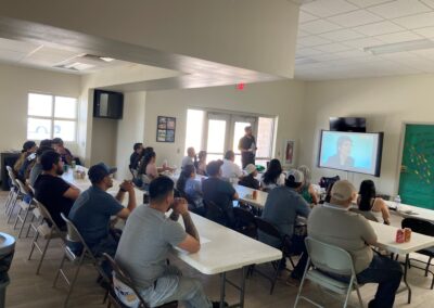Fathers and family members sit at long tables watching a presentation on a large screen in a community room.