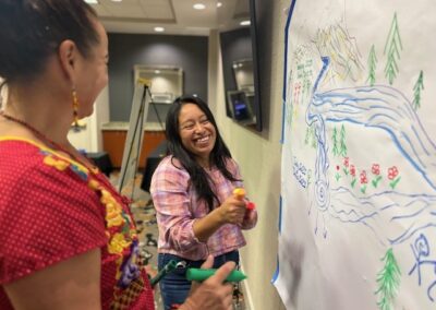 Two family leaders laugh together beside a large hand-drawn mural of a river, mountains, and forest.