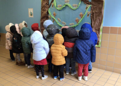 A group of preschoolers in winter coats stand together looking up at a classroom bulletin board.